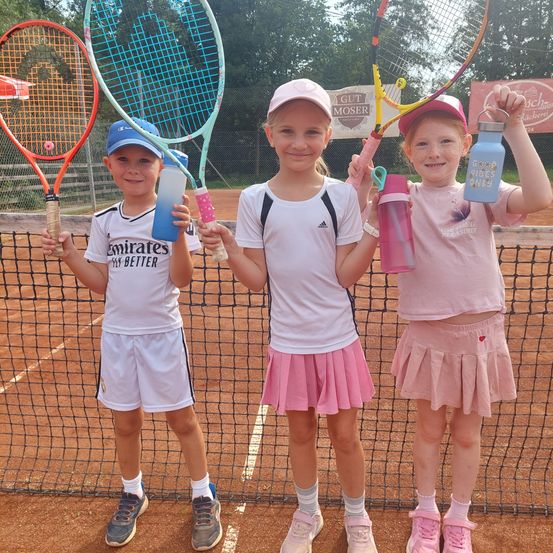 Drei junge Mädchen stehen auf einem Tennisplatz und halten Tennisschläger und Wasserflaschen in den Händen. Sie sind in passenden Outfits gekleidet und scheinen für ein Foto zu posieren.