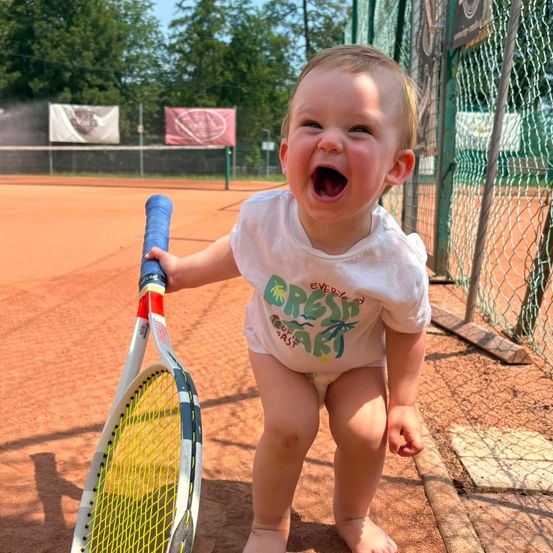 Ein Kleinkind in einem weißen T-Shirt mit grünem Aufdruck und Windel hält einen Tennisschläger auf einem Tennisplatz.