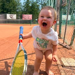 Ein Kleinkind in einem weißen T-Shirt mit grünem Aufdruck und Windel hält einen Tennisschläger auf einem Tennisplatz.