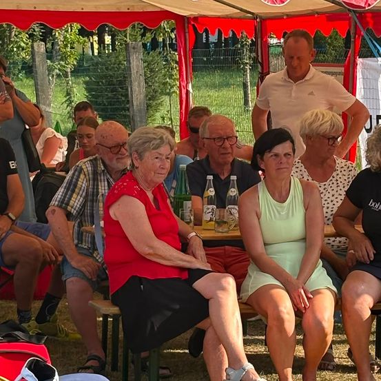 Eine Gruppe älterer Menschen sitzt in einem Zelt, einige tragen Brillen und Sandalen, mit einem Mann, der hinter ihnen steht. Flaschen und Gläser stehen auf dem Tisch.