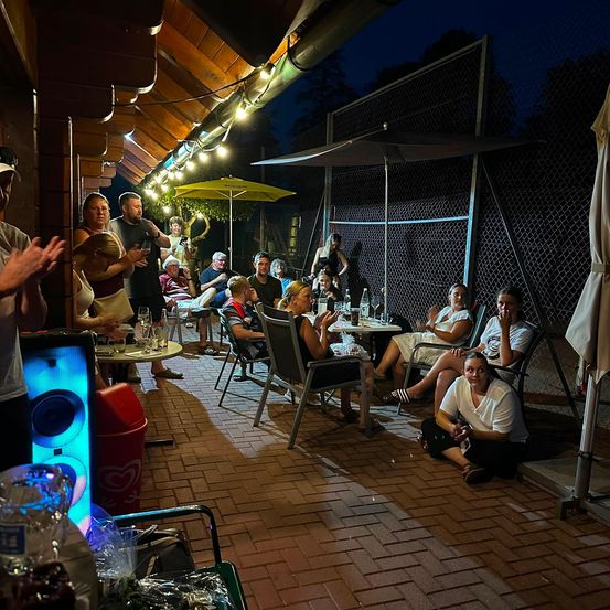 Eine Gruppe von Menschen sitzt und steht um Tische in einem Outdoor-Restaurant bei Nacht, mit Schirmen und Lichtern für Ambiente.