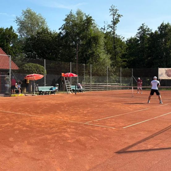 Zwei Tennisspieler spielen auf einem Sandplatz mit Netz, umgeben von einem Zaun und Bäumen. Zuschauer beobachten von den Seitenlinien.