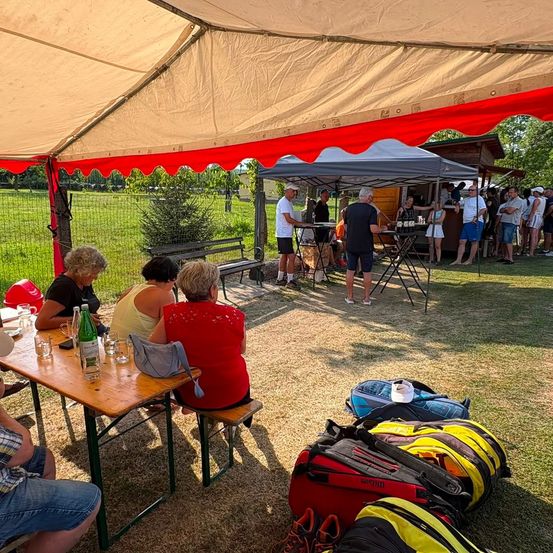 Eine Gruppe von Menschen versammelt sich unter einem Zelt in einem Park. Einige sitzen an einem Tisch, während andere in der Nähe eines Essensstandes stehen. Es gibt Taschen und Flaschen auf dem Boden.