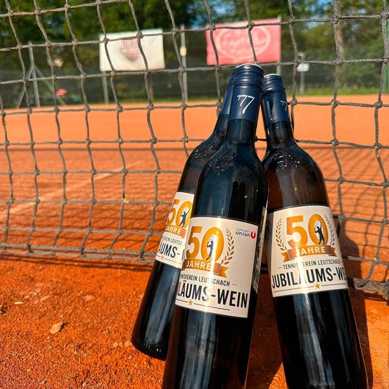 Drei Flaschen 50 Jahre Jubiläums-Wein stehen auf einem Tennisplatz, mit einem Netz im Hintergrund.
