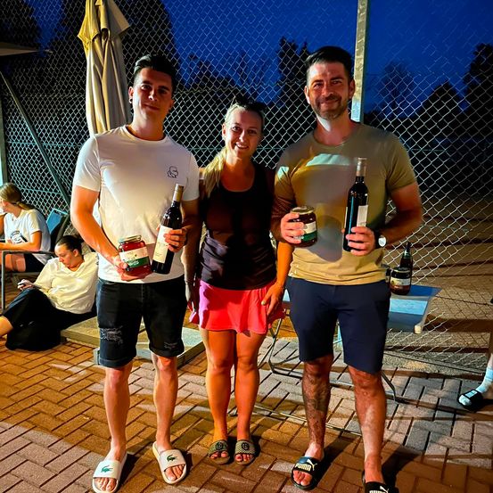 Drei Menschen stehen zusammen und halten Flaschen Wein in der Hand, einer von ihnen trägt ein weißes T-Shirt, ein anderer ein schwarzes T-Shirt und rote Shorts, und der dritte ein beiges T-Shirt. Sie stehen draußen in der Nähe eines Zauns.