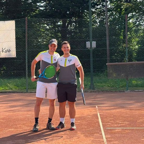 Zwei Männer stehen auf einem Tennisplatz, einer hält eine Tennisschläger in der Hand, beide lächeln für die Kamera. Hinter ihnen ist ein Zaun mit einem Banner zu sehen, auf dem 'Karo' steht.