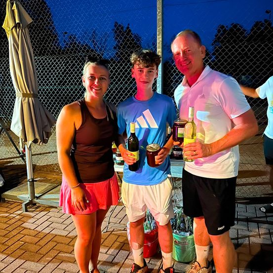 Drei Personen stehen bei Nacht zusammen und lächeln für ein Foto. Eine Frau in einem braunen Tanktop und einer pinken Rock, ein Junge in einem blauen Adidas-Shirt und ein Mann in einem weißen Poloshirt. Sie halten Flaschen Wein und Dosengetränke. Hinter ihnen steht ein Zaun und ein geschlossener Regenschirm.