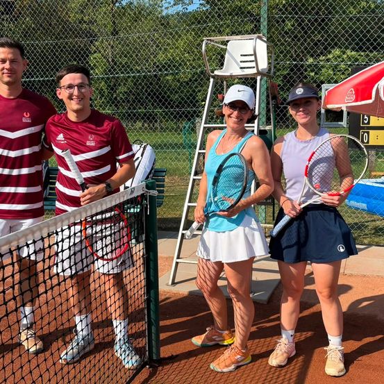 Vier Personen, zwei Männer und zwei Frauen, stehen auf einem Tennisplatz und halten Tennisschläger. Die Frauen tragen weiße und blaue Röcke, während die Männer rote und weiße Trikots tragen. Ein Netz und ein Tennisstuhl stehen hinter ihnen.
