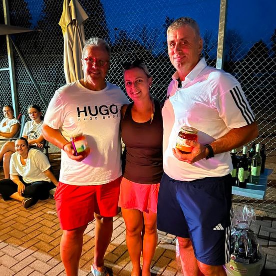 Drei Menschen stehen zusammen, halten Gläser mit Essen und lächeln für ein Foto. Hinter ihnen ist ein Zaun und ein Tisch mit Flaschen und einem Korb zu sehen. Im Hintergrund sitzt eine Gruppe von Menschen auf dem Boden.