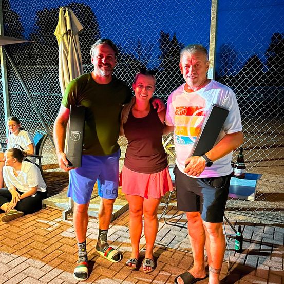 Drei Menschen stehen zusammen und halten Trophäen, lächeln für ein Foto bei Nacht. Ein Zaun und Bäume sind im Hintergrund.
