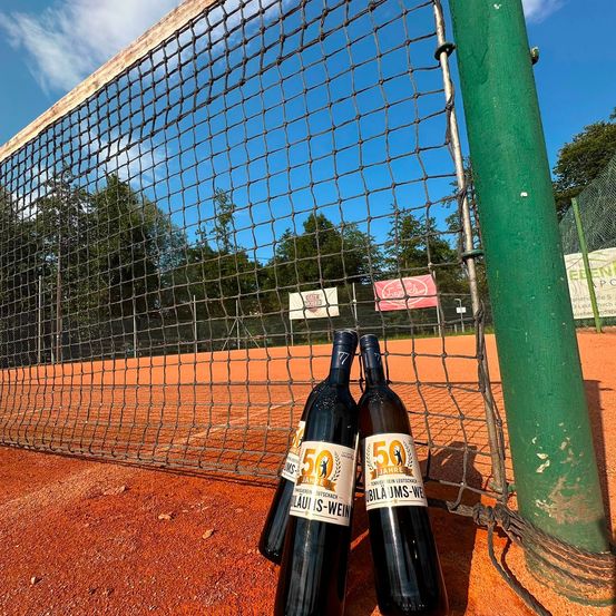 Drei Weinflaschen stehen auf einem Tennisplatz, mit einem Netz im Hintergrund und einem klaren blauen Himmel darüber.