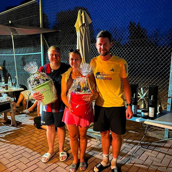 Drei Menschen stehen lächelnd nebeneinander mit Geschenkkörben in der Hand. Der Mann auf der linken Seite hält einen grünen Korb, die Frau in der Mitte einen roten und der Mann auf der rechten Seite trägt ein gelbes T-Shirt. Dahinter sind ein Zaun und ein Tisch mit Flaschen und einem geschlossenen Regenschirm zu sehen.