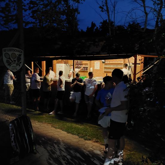 Eine Gruppe von Menschen steht nachts vor einer kleinen Bar. Die Bar hat ein Schild mit der Aufschrift 'SU LEUTSCHACK 1976'. Einige Leute stehen um eine Theke herum und halten Getränke in den Händen. Ein Tennisschläger ist auf dem Schild zu sehen.