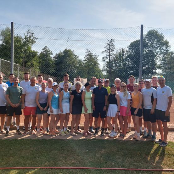 Eine Gruppe von Menschen in Sportkleidung posiert für ein Foto auf einem Tennisplatz. Im Hintergrund befinden sich ein Zaun und Bäume.