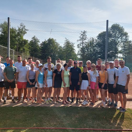 Eine Gruppe von Menschen in Sportkleidung, die vor einem Tennisplatz mit Zaun und Bäumen im Hintergrund stehen.
