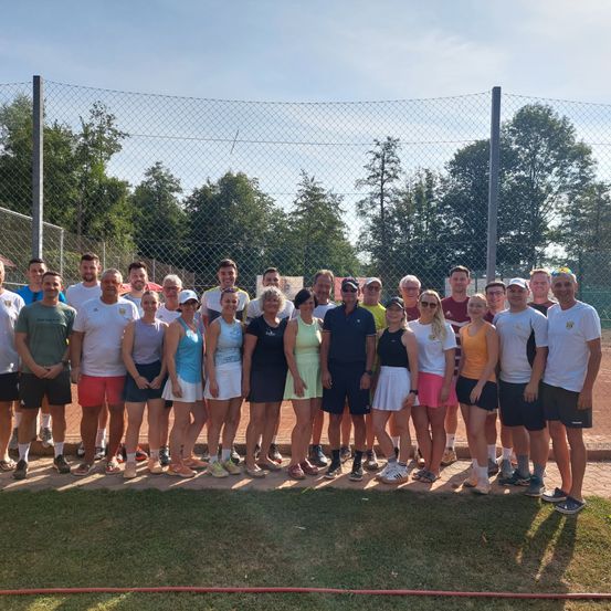 Eine Gruppe von Tennisspielern, sowohl Männer als auch Frauen, in verschiedenen farbigen Outfits gekleidet, stehen vor einem Gitterzaun an einem sonnigen Tag.