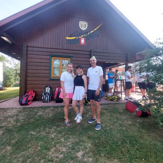 Drei Personen stehen vor einem Holzgebäude mit Logo und Tennisausrüstung auf dem Boden.