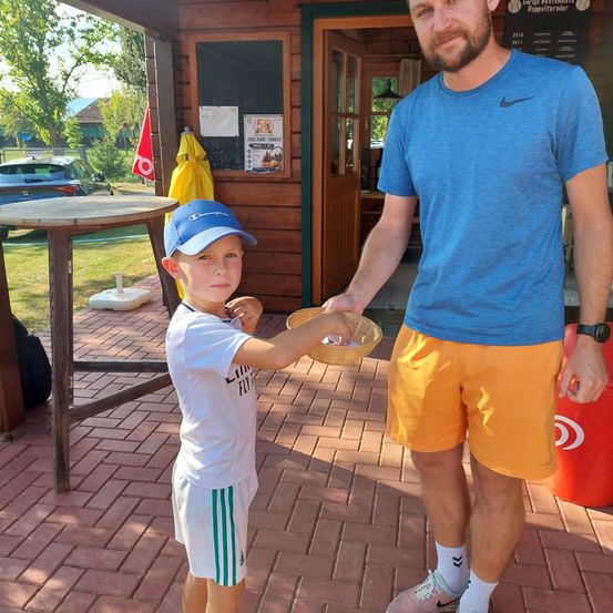Ein junger Junge in einer blauen Kappe und einem weißen Shirt hält einen Korb, während er neben einem Mann in einem blauen Shirt und orangefarbenen Shorts steht, beide vor einem hölzernen Gebäude.