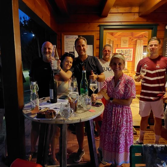 Eine Gruppe von Menschen versammelt sich um einen Tisch in einer Holzhütte, lächelt und hält Weingläser in den Händen. Eine Frau in einem rosa Kleid stößt mit einem Mann in einem schwarzen Shirt an. Flaschen und Gläser stehen auf dem Tisch, und ein Mann in einem roten Shirt steht in der Nähe.