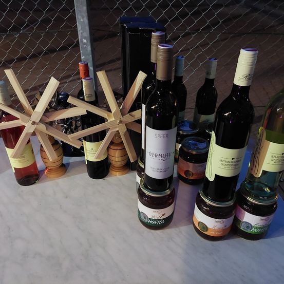 Eine Sammlung von Weinflaschen, Marmeladengläsern und einer hölzernen Windmühle auf einem Marmortisch. Die Flaschen sind mit Namen wie Speer, Weissburgunder und anderen beschriftet. Der Tisch steht vor einem Zaun.