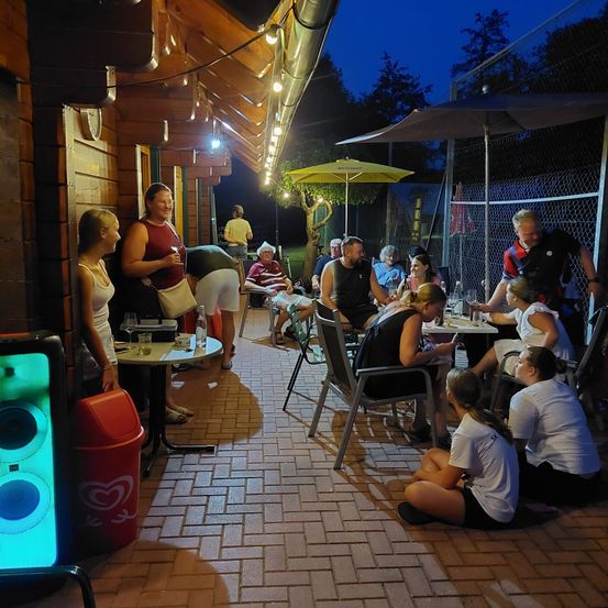 Eine Gruppe von Menschen versammelt sich bei Nacht auf einer Terrasse um Tische, einige sitzen und andere stehen, eine Frau hält ein Glas und ein Mann steht in der Nähe.
