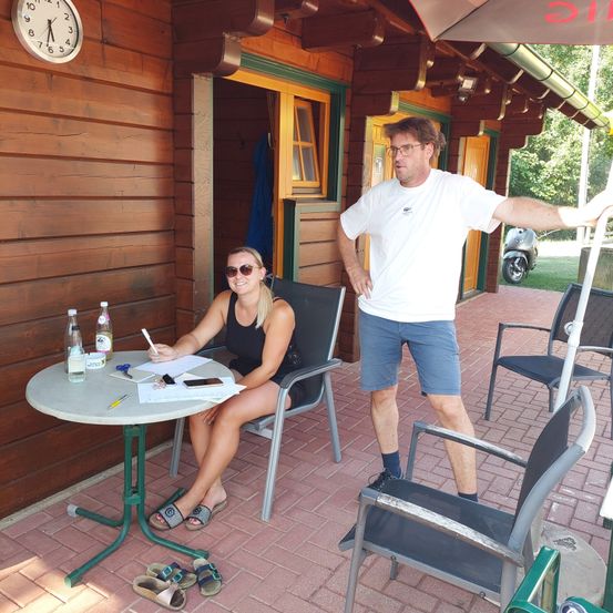 Ein Mann und eine Frau stehen auf einer Terrasse. Die Frau sitzt an einem Tisch und schreibt auf ein Stück Papier. Der Mann steht hinter ihr und lehnt sich an ein Geländer.