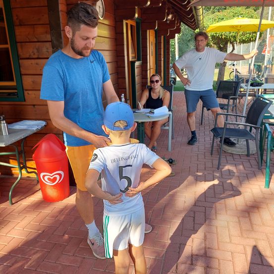 Ein Mann justiert das Trikot eines jungen Jungen, der ein Trikot mit der Nummer 5 trägt. Hinter ihnen sitzt eine Frau am Tisch, und ein Mann steht mit den Händen auf den Hüften.