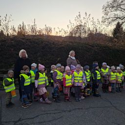 Eine Gruppe von Kindern mit gelben Weste und Stiefeln stehen in einer Reihe, begleitet von zwei Frauen, mit einem Busch im Hintergrund und einer untergehenden Sonne.
