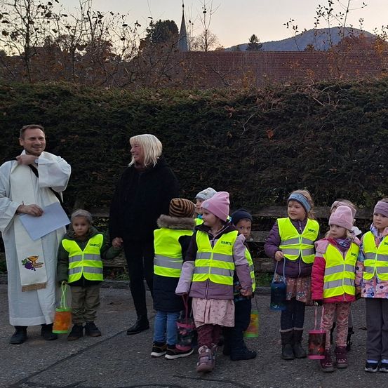 Eine Gruppe von Kindern und zwei Erwachsenen steht draußen. Die Kinder tragen neongelbe Westen und tragen orange Taschen. Der Mann hält ein Papier. Ein Busch und Gebäude sind im Hintergrund.