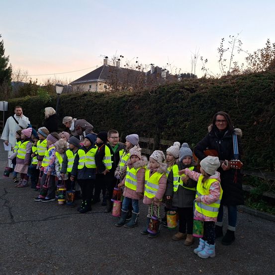 Eine Gruppe von Kindern in gelben Westen steht auf einer Straße mit einer Frau, die Gitarre spielt. Ein Haus ist hinter ihnen.