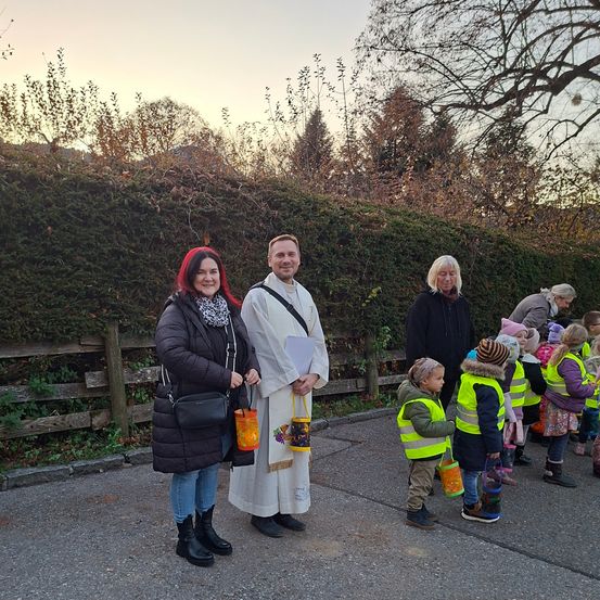 Eine Gruppe von Menschen steht vor einer Hecke, darunter ein Mann in Weiß, eine Frau mit roten Haaren und mehrere Kinder, die reflektierende Westen tragen.