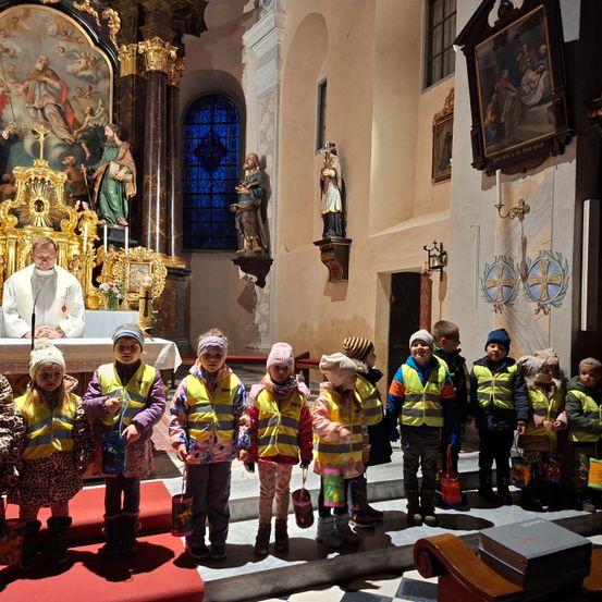 Eine Gruppe von Kindern in gelben Westen steht in einer Kirche und schaut den Priester am Altar an.
