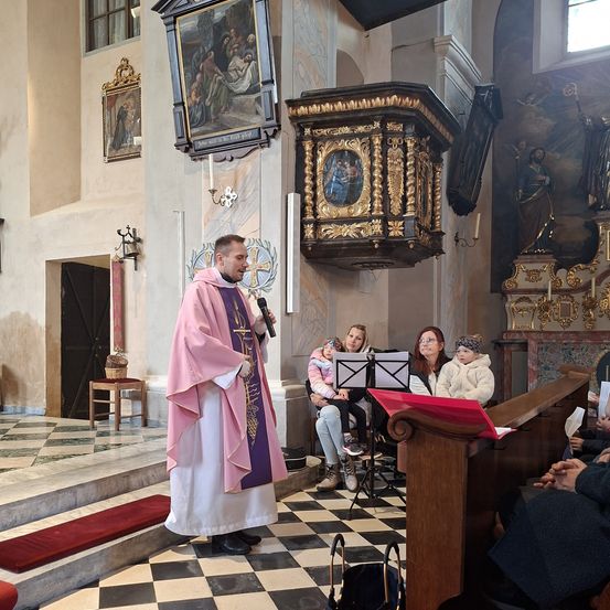 Ein Priester steht in einer Kirche und spricht in ein Mikrofon. Leute sitzen auf den Bänken, einige mit Babys. Kunstwerke und religiöse Symbole zieren die Wände.