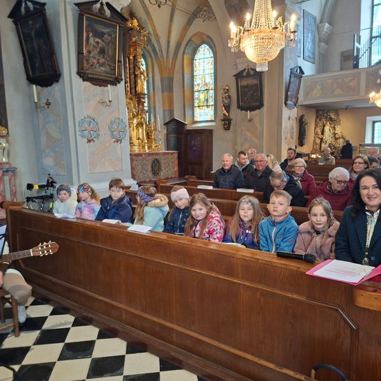 Eine Gruppe von Kindern und Erwachsenen sitzt in hölzernen Kirchenbänken. Eine Person spielt links eine Gitarre.