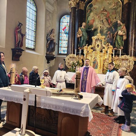 Eine Gruppe von Menschen steht in einer Kirche, einige in weißen Gewändern, vor einem Altar. Ein Priester hält einen Kelch. Statuen und Buntglasfenster schmücken den Hintergrund.