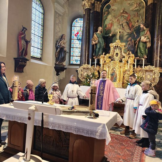 Eine Gruppe von Menschen steht in einer Kirche, einige in weißen Gewändern, vor einem Altar. Ein Priester hält einen Kelch. Statuen und Buntglasfenster schmücken den Hintergrund.