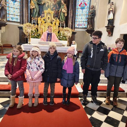 Eine Gruppe von Kindern und Jugendlichen steht in einer Kirche, mit einem Priester am Altar hinter ihnen. Die Kinder tragen Winterkleidung und Stiefel.