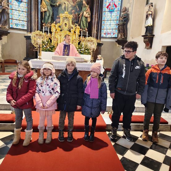 Eine Gruppe von Kindern und Jugendlichen steht in einer Kirche, mit einem Priester am Altar hinter ihnen. Die Kinder tragen Winterkleidung und Stiefel.