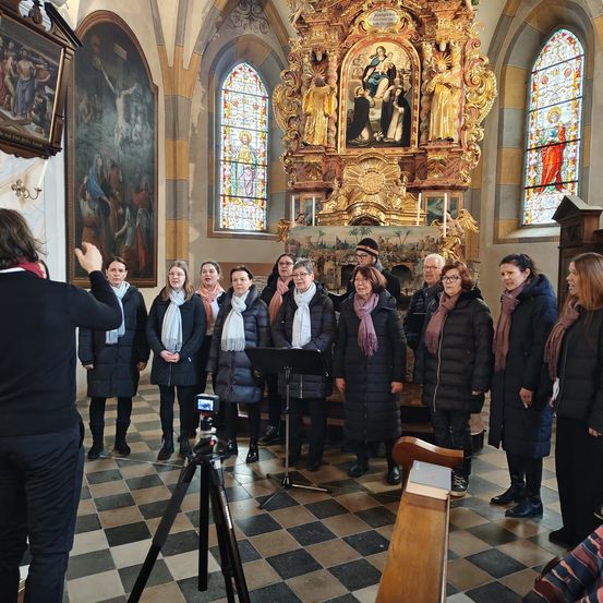 Eine Gruppe von Frauen in Winterkleidung steht in einer Kirche, möglicherweise singend. Sie stehen vor einem goldenen Altar mit Gemälden.