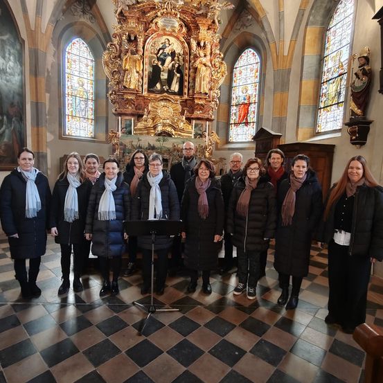 Eine Gruppe von Menschen in Wintermänteln, Schals und Stiefeln posiert für ein Foto in einer Kirche. Sie stehen vor einem Notenständer, und hinter ihnen befindet sich ein kunstvoller Altar mit einer Statue und Buntglasfenstern.