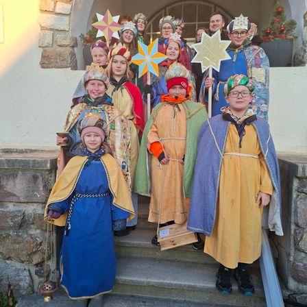 Eine Gruppe von Kindern in Kostümen steht auf den Stufen eines Gebäudes und hält Requisiten wie Sterne und Kronen.