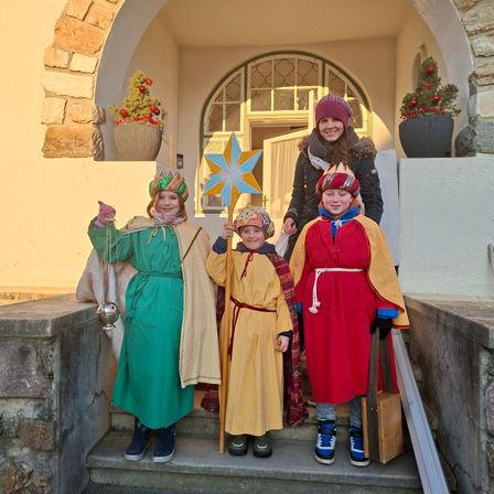 Drei Kinder in mittelalterlichen Kostümen stehen auf den Stufen eines Gebäudes mit einer Frau, die einen Stern hält. Die Kinder halten eine Glocke, einen Stab und ein Buch. Dahinter befindet sich ein Bogen mit Topfpflanzen.