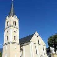 Eine weiße Kirche mit einem hohen Turm und Uhren, gelegen unter einem klaren blauen Himmel, mit mehreren Rundbogenfenstern und einer Statue davor.