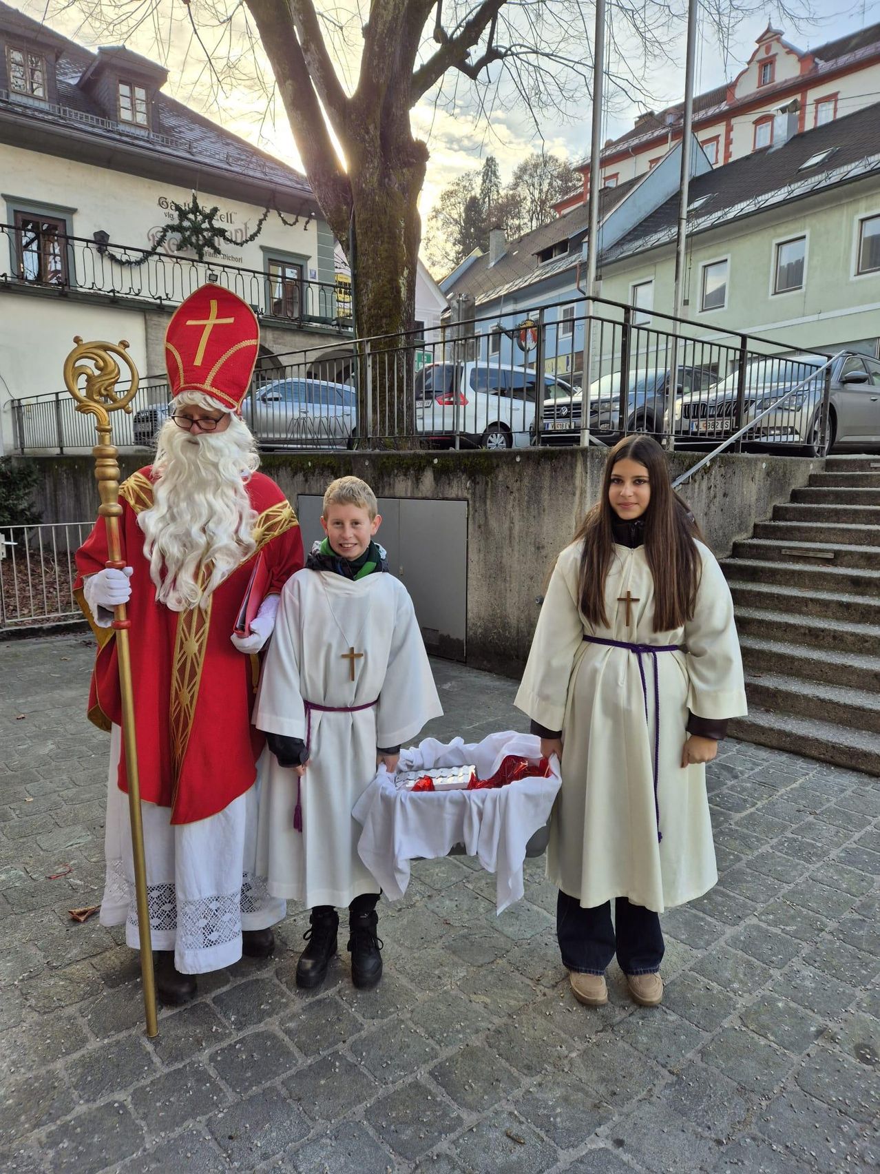 Ein Mann in der Kleidung des Heiligen Nikolaus steht mit zwei Kindern in religiösen Gewändern. Der Junge hält einen Korb. Sie stehen vor einem Gebäude mit Balkon und Treppen. Autos sind in der Nähe geparkt.