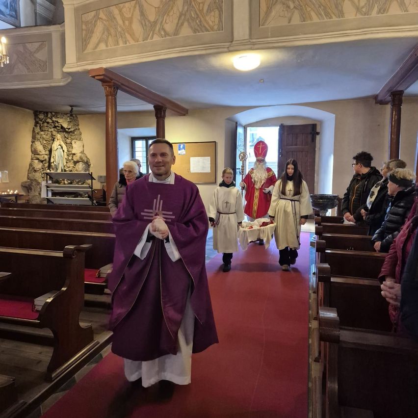 Ein Priester in violetten Gewändern geht einen roten Teppich in einer Kirche mit einer Statue der Jungfrau Maria und zusehenden Leuten entlang.