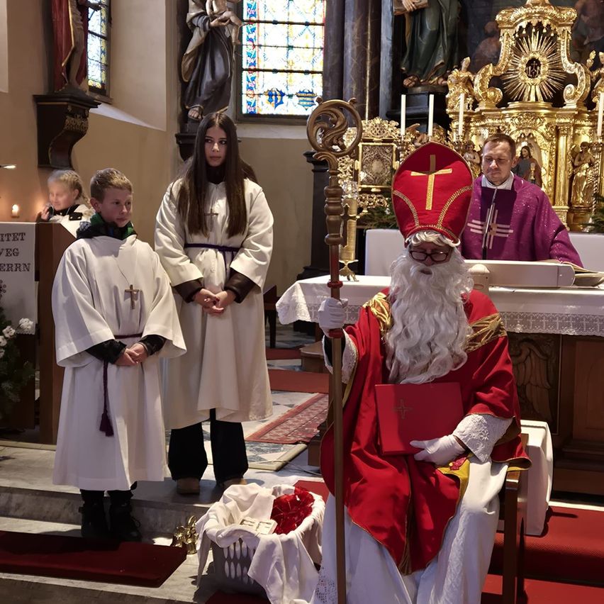 In einer Kirche sitzt ein als Weihnachtsmann verkleideter Mann mit einem Stab auf einem Stuhl. Ein Mann in einer Priesterrobe steht hinter einem Tisch. Zwei Kinder in weißen Roben stehen vor ihm.