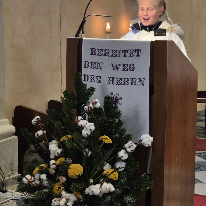 Ein junges Mädchen steht hinter einem Podium mit einer dekorativen Pflanze davor. Das Podium hat ein Schild mit der Aufschrift 'Bereitet den Weg des Herrn.' Hinter ihr steht eine Kerze auf einem Ständer.
