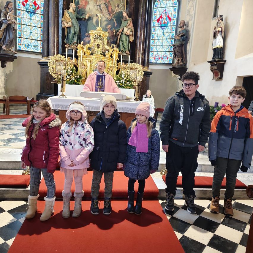 Eine Gruppe von Kindern steht vor einem Priester am Altar einer Kirche, mit Statuen und Buntglasfenstern im Hintergrund.