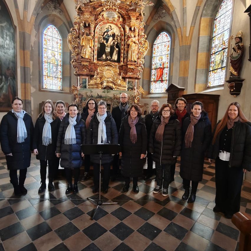 Ein Chor steht in einer Kirche mit einem goldenen Altarbild hinter ihnen. Sie sind alle in Wintermänteln und Schals gekleidet.