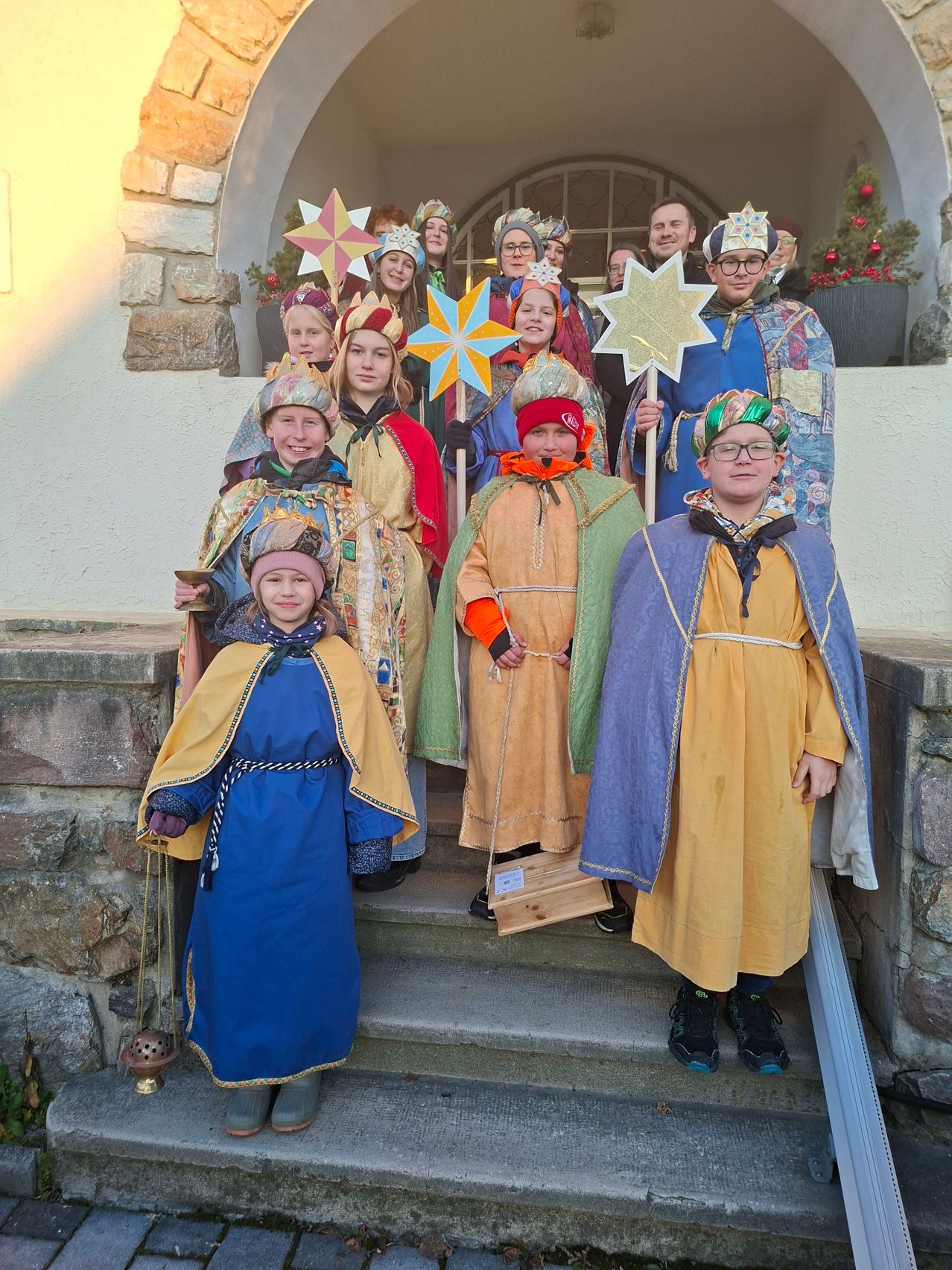 Eine Gruppe von Kindern in bunten Kostümen, möglicherweise für ein Weihnachtsevent, steht auf den Stufen eines Gebäudes. Sie halten einen Stern und eine Laterne.